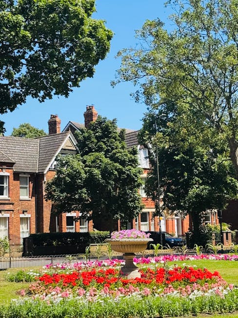 A well-maintained front garden in Raphael Park featuring neatly trimmed, rounded bushes and small conifer trees arranged along a curved, paved pathway bordered by dark soil. The lush grass lawn is vib