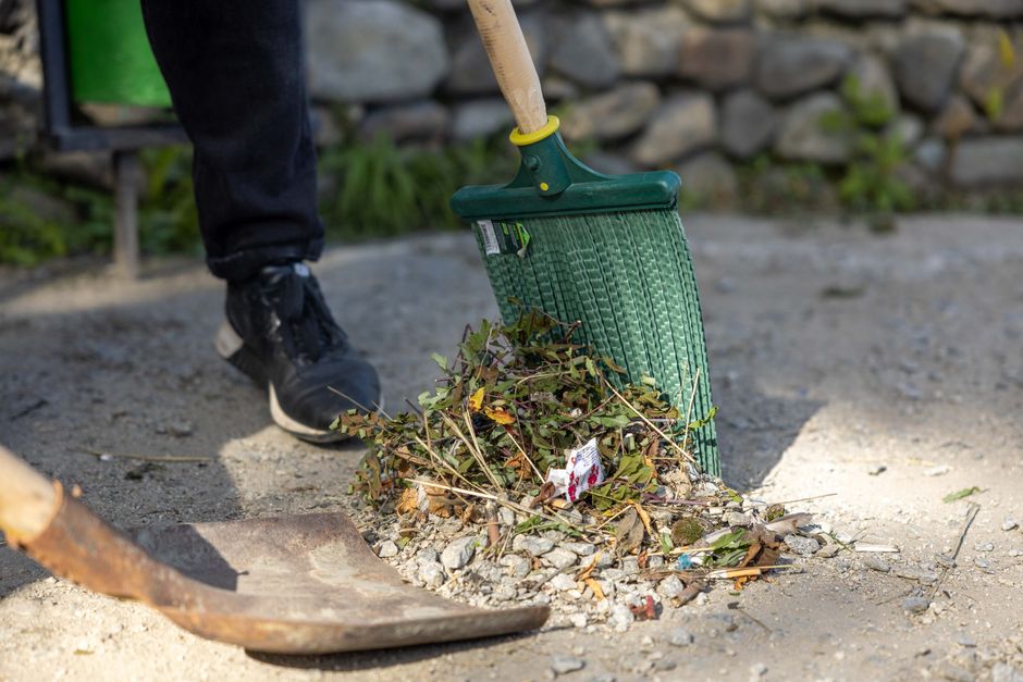 A person wearing black sneakers and dark clothing is using a green hand rake to clear garden debris, including leaves, small branches, and gravel, onto a pile on a garden pathway. The pathway appears 